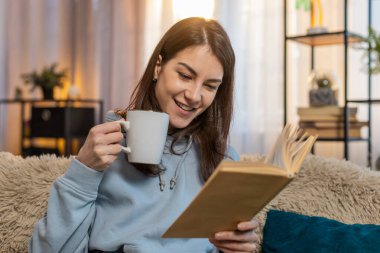 Rahat koltukta bir fincan sıcak çikolata, kakao ya da çay içerken ilginç bir kitap okuyarak rahatlayan genç kadın edebiyatın tadını çıkarıyor. Evdeki oturma odasında sıcak bir battaniyeye sarılı gülümseyen kız..