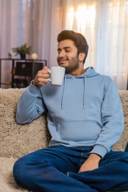 Gülümseyen Hintli adam evde sıcak kahve ya da bitki çayı içiyor. Hindu genç adam moladan zevk alıyor, sabahları kanepede oturuyor, düşünceli bir şekilde uzaktan bakıyor ve dinleniyor.