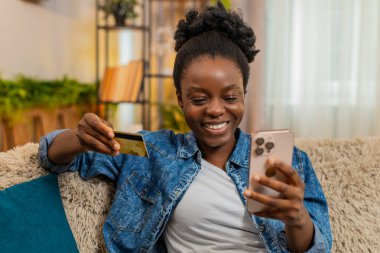 Afro-Amerikalı genç bir kadın internet alımını mutlu bir şekilde akıllı telefon ve kredi kartıyla tamamlıyor. Rahatlıkla gülümsüyor ve tatmin oluyor. Siyah kız evde rahat bir koltukta alışveriş yapmayı sever..