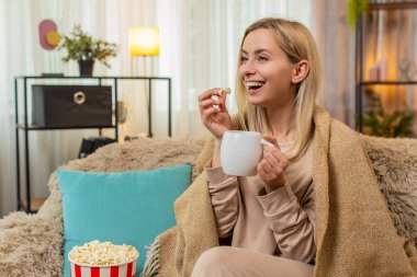 Genç, yetişkin bir kadın sıcak bir battaniyeye sarılmış, sıcak bir çay tutuyor ve sakin bir şekilde diziyi seyrediyor. Sarışın kız düşünceli görünüyor, sıcak içki ve konforla huzurlu bir anın tadını çıkarıyor..