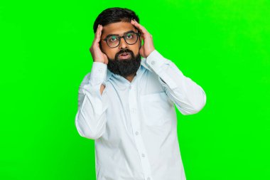 Hintli adam gergin yüzüyle parmaklarını şakaklarına bastırır, baş ağrısı taklidi yapar ya da stresli duygularla zihinsel aşırı yüklenme yapar. Yeşil krom anahtar arka plandaki Arap adam basınç ve acı reaksiyonu gösteriyor.