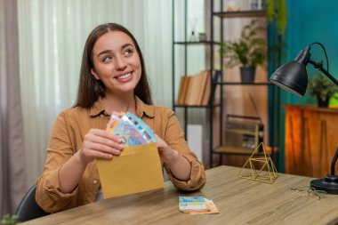 Evdeki genç kadın avroya ait nakit paraları sayıyor, gelecekteki satın alma planlarının bir kısmını zarfa atıyor. Beyaz kız sessizce gülümsüyor. Para ve kişisel finansal hedefler konusunda kendini güvende hissediyor..