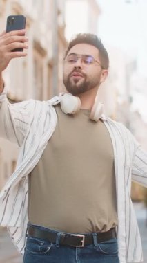 Beyaz erkek blogcu akıllı telefondan selfie çekiyor, abonelerle çevrimiçi video görüşmesi yapıyor, sosyal medya vlog 'u için hikayeler kaydediyor. Şehir caddesinde duran mutlu adam.