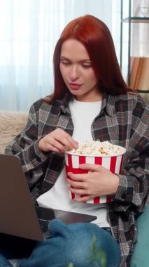 Evde dizüstü bilgisayarda film izleyen, patlamış mısır yiyen ve neşe ve heyecanla gülen genç bir kadın. Kanepede oturan beyaz saçlı kız duygusal sahnenin tadını çıkarıyor, eğlence dolu bir gece geçiriyor.