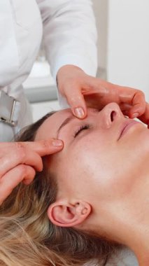Beyaz bir kadın kozmetoloji kliniğindeki kaş bölgesindeki yüz masajı sırasında spa kanepesinde dinleniyor. Kozmetik uzmanı sakin olmak, cilt bakımı ve cilt tazeleme için nazik dokunuşlar kullanır.