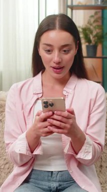 Evdeki genç kadın akıllı telefon mesajı okuyor ve yarışmayı yüksek sesle mutlu bir şekilde kazanmasını kutluyor. Kafkasyalı kız telefonu sımsıkı tutarak neşeli bir şok geçiriyor ve samimi bir odada gülüyor..