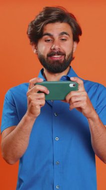 Endişeli komik bağımlı adam cep telefonuyla yarış oyunları oynuyor. Turuncu stüdyo arka planında izole edilmiş sürücü simülatörlü akıllı telefon uygulaması kullanan mutlu bir orta doğulu.