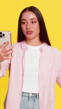 Genç bir kadın akıllı telefondan yapılan bir video görüşmesi sırasında enerjik bir şekilde el sallıyor neşeli ruh haliyle iletişim kuruyor. Sarı arka plandaki kız çevrimiçi animasyon konuşmalarında heyecan verici bir ilişki gösteriyor.