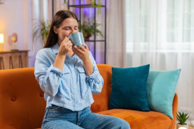 Oldukça mutlu gülümseyen Kafkasyalı genç bir kadın sabahın köründe sıcak kahve ya da bitki çayı içerek portakal rengi kanepede oturuyor. Oturma odasında tek başına rahatlamak isteyen bir kız.