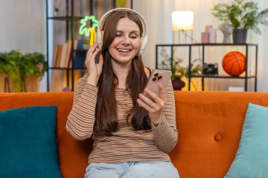 Evde kablosuz kulaklık kullanan genç bir kadın mesaj atarken başını sallayıp gülümsemeye başlıyor. Kanepede oturan beyaz bir kız elinde telefonuyla mesajlara cevap veriyor. Yumuşak bir gülümsemeyle kafa sallıyor ve cevap veriyor.