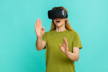 Orta yaşlı kadın VR kulaklık takıyor ve dikkatlice etrafa bakıyor, meraklı odaklı yüzüyle dijital çevreyi keşfediyor. Mavi arka planda izole edilmiş kız sanal dünyaya ilgi göstererek tepki veriyor