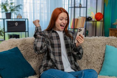 Evde akıllı telefondan güzel haberler okuyan, gülümseyen ve heyecanla el kaldıran genç bir kadın. Kafkasyalı kızıl saçlı kız kanepeye oturmuş neşeli bir başarıyı kutluyor, mutlu, olumlu duygusal bir zafer yaşıyor.