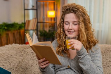 Hamile genç bir kadın evde kanepede oturmuş taze portakal suyu yudumluyor ve rahat bir ifadeyle hamilelik kitabı okuyor. Kız, gelecekteki anneliği ve nazik yaşam tarzını yansıtan huzurlu bir anın tadını çıkarıyor.