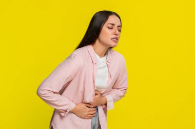 Karnını ovuşturan genç bir kadın. Acı dolu bir ifadeyle öne doğru eğiliyor. Karın ağrısından muzdarip olduğu çok açık. Sarı arka plandaki kız mide ağrısı ve sindirim sorunu belirtileri gösteriyor..