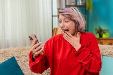 Kanepede zaferi duygusal olarak kutlayan genç bir kadın akıllı telefon ekranına bakarak neşeli bir heyecan yaşıyor. Kafkasyalı kız şaşırdı. Mobil oyunu kazanınca hayrete düştü. El sallıyor.