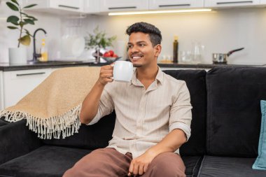 Evdeki Hintli adam çay bardağını tutuyor sıcak elleri tutuyor küçük bir yudum alıyor rahatlatıyor ve güne barışla başlıyor. Kanepedeki Arap adam sıcak içecekten hoşlanıyor. Nefes alıyor ve zihnini çaya hazırlıyor. Sakin ol.