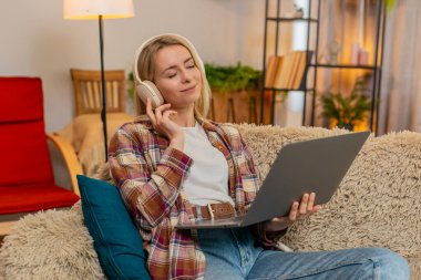 Genç yetişkin bir kadın evdeki kanepede otururken bilgisayarında en sevdiği müziği kablosuz kulaklıkla dinliyor. Mutlu kız gülümsüyor ve neşeyle ritme göre hareket ediyor, pozitif enerji ve rahat ruh hali gösteriyor..
