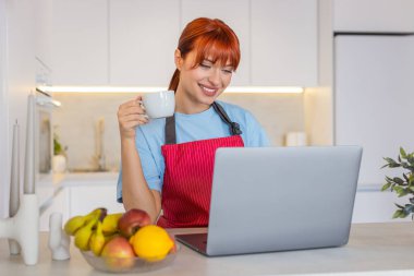 Evde oturan genç bir kadın dizüstü bilgisayarla e-postalarına cevap veriyor ve kahveden bir yudum alıyor. Mutfaktaki kızıl saçlı kız, gelen kutusuna cevap veriyor. Müşteriler için taslak notlar. İşine odaklan..