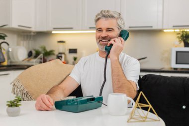 Evdeki orta yaşlı bir adam telefon hattını kaldırıyor nostaljiyle gülümsüyor ve koltuktaki masada sıcak bir merhaba diyor. Olgun adam cep telefonunu yakın paylaşıyor. Nazik sözler söylüyor. Sıcak bir ton ve kısa konuşmadan hoşlanıyor.
