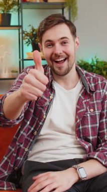 Heyecanlı beyaz adamın tasvip edici bir şekilde kameraya bakan portresi, işaret memnuniyeti, iyi haber, olumlu geri bildirim gibi. Evdeki oturma odasındaki kanepede oturan mutlu genç adam.