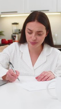 Evdeki genç bir kadın, yeni bir kopya basmak için dizüstü bilgisayarının yanına sayfa fırlatmış. Kanepede oturan beyaz kız üzgün görünüyor. Yeni fatura çekleri hazırlıyor.