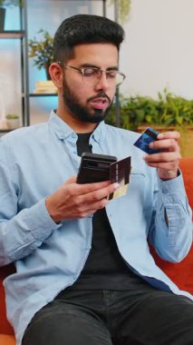 İnternetten alışveriş yapan mutlu genç Hintli adam. Gülümseyen İspanyol adam elinde bankacılık kredi kartı tutuyor evdeki koltukta mobil uygulamanın doğrulama sürecinde yer alıyor..