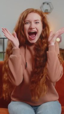 Mutlu genç bir kadın TV izlerken bağırıyor, başarıyı kutluyor, kazanıyor, gol başarısı iyi haber, piyango şansı, oturma odasında koltukta oturan zafer. Yetişkin kız evde çifte onay gösteriyor..