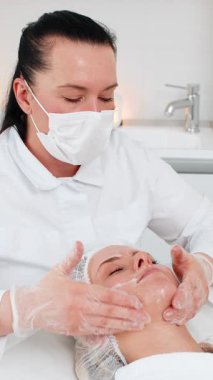Yüz masajı soyuluyor. Güzellik uzmanı kozmetik uzmanı cilt bakımı ve gençleştirme için kozmetoloji kliniğinde orta yaşlı kadın yüzüne krem ve serum uyguladı.