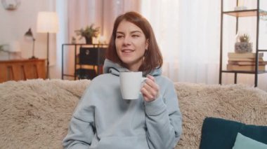 Mutlu genç bir kadın, içecek kokusu alıyor sıcak kahve içiyor, mola veriyor, dinleniyor. Gülümseyen beyaz kız aroma çayını tek başına içmeyi sever. Oturma odasındaki kanepede oturur..