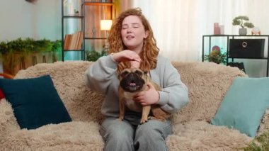 Mutlu Kafkasyalı kızıl saçlı kadın pug dog 'u koltukta nazikçe okşuyor, sıcakkanlılıkla ve yumuşak duygularla gülümsüyor. Evcil hayvan yakınımızda kalır, temastan zevk alır ve evde samimi bir dostluk içinde birlikte dinlenir.