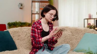 Mutlu kadın evdeki koltukta akıllı telefona bakıyor, iyi haberler aldıktan sonra sevinçle tepki veriyor ve piyango kazanıyor. Kız online piyangoyu zafer jesti, görünür mutluluk ve memnuniyet ile kutluyor.