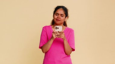 Hintli kadın gülümsüyor, akıllı telefon tutuyor, neşeli mesajlar atıyor, çevrimiçi sohbet ediyor. Bej arka plandaki kız pozitif iletişim sosyal medya etkileşiminde aktif mutluluk sohbeti gösteriyor