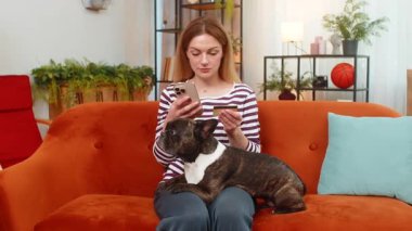 Evde buldogu olan orta yaşlı mutlu bir kadın akıllı telefon uygulamasında köpek maması sipariş ediyor. Kredi kartı ödemeleri, iyi niyet göstergesi. Kanepedeki kız, sakin gözlerle bekler. Rahatlık sevinç verir.