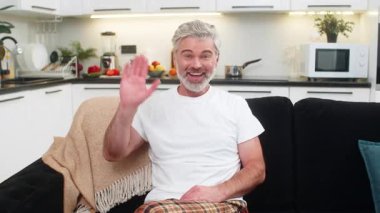 Evde kameraya el sallayan orta yaşlı bir adam merhaba diyor ve arkadaş canlısı gülümsemelerle çevrimiçi iletişim kurmaya başlıyor. Kanepedeki olgun adam lenslere bakar kısa selam verir sıcak hava konuşur açık ton sakin nefes al istikrarlı.