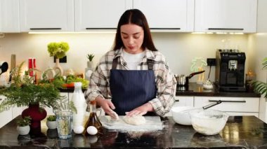 Genç bir kadın üzerine un serper ve hamur ezer. Süt, yumurta ve yağın yanında önlük giyer. Evde pişirme işlemi için pizza tabanı ya da ekmek hazırlıyorum. Temiz mutfak konsepti ve sakin bir sabah rutini..