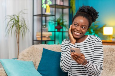 Afrikalı Amerikalı kadın sosyal medya profilini güncelliyor. Evdeki kanepede oturmak, fotoğraf çekmek, tasvirler yazmak, mime gülmek. Rahatlamış ruh hali, kapalı mekanda gelişigüzel etkileşim. Çevrimiçi zaman geçirmek eğlenceli