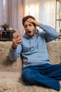 Akıllı telefon kullanan, negatif mesaj okuyan, oturma odasında kanepede oturmaktan rahatsız olan üzgün genç adam şok oldu. Alet sorunu olan Hintli adam üzgün görünüyor, telefonu tutuyor ve kötü habere tepki veriyor..