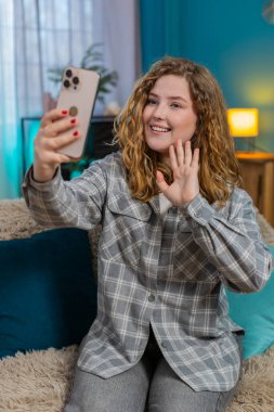 Genç bir kadın akıllı telefonuyla arkadaşını sevinçle karşılıyor ve mutlu haberi paylaşıyor. Kafkasyalı kızıl kız kanepede dinleniyor, neşeli bir şekilde gülümsüyor, pozitif etkileşim içinde..