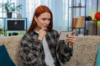 Evde kulaklık takmış akıllı telefondan video izleyen genç bir kadın. Kanepedeki beyaz saçlı kız eğlenceli sosyal medya içeriğinin keyfini çıkarıyor, neşe ve neşeyle tepki veriyor.