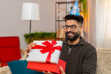 Evdeki Hintli adam mutlu bir şekilde doğum günü hediyesini açarken koltukta gülümseyerek ve heyecanla oturuyor. Arap adam şaşırmış ve neşeli görünüyor. Beklenmedik bir hediye ve özel bir kutlama anı karşısında heyecanlanıyor.