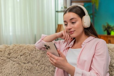 Genç bir kadın evdeki kanepede kablosuz kulaklık takarak oturuyor ve akıllı telefonuyla podcast dinliyor ve notlar yazıyor. Beyaz kız yeni bilgilerden memnun ve öğrenirken ritim tutuyor..