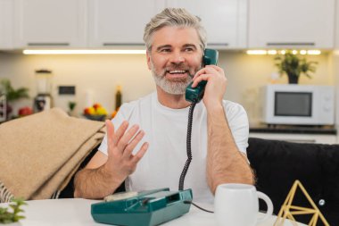 Evdeki orta yaşlı bir adam telefon hattını kaldırıyor nostaljiyle gülümsüyor ve koltuktaki masada sıcak bir merhaba diyor. Olgun adam cep telefonunu yakın paylaşıyor. Nazik sözler söylüyor. Sıcak bir ton ve kısa konuşmadan hoşlanıyor.