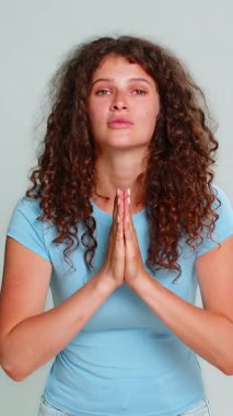 Genç bir kadın esprili bir şekilde el çırpıyor dua ediyor gibi yapıyor, umut dolu, neşeli bir ifadeyle yardım dileniyor ya da yardım dileniyor. Gri arkaplanda duygusal jestler yapan bir kız şakacı ve komik bir şekilde rica ediyor..