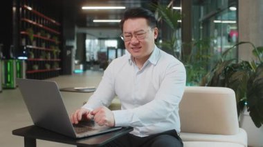 Gergin Çinli yetişkin iş adamı dizüstü bilgisayar kullanıyor ve modern ofis lobisinde çalışırken kalp krizi geçirme riski olan bir göğsü tutmaktan muzdarip. İş adamı gömlekli..