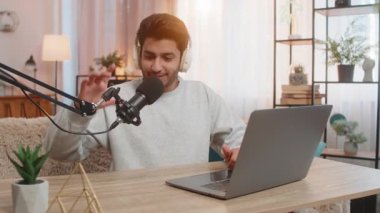 Genç Hintli adam kulaklık ve stüdyo mikrofonuyla laptop ve webcam 'e bakıyor, vlog kaydediyor, online akış yapıyor ve evdeki oturma odasında otururken aboneleri selamlıyor..