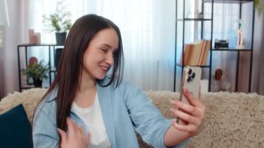 Evde oturan genç bir kadın video aracılığıyla flört ediyor, göz kırpıyor, neşeli bir şekilde saçlarını düzeltiyor. Kafkasyalı güzel kız romantik ruh hali, olumlu duygular, mutlu etkileşim