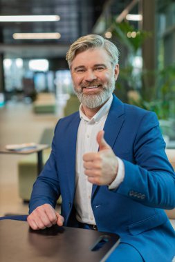 Gibi. Takım elbiseli mutlu, orta yaşlı bir işadamı kameraya bakıp olumlu bir işaret veriyor. Modern ofis lobisinde oturmuş olumlu bir izlenim veriyor. İş insanları.