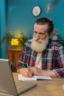 İnternette dizüstü bilgisayarda çalışan kıdemli işadamı ofis çalışma masasındaki planlı not defterine notlar yazıyor. Profesyonel serbest çalışan büyükbaba. Uzaktan eğitim, eğitim, öğrenme dersleri. Dikey