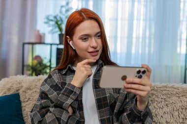 Evde kulaklık takmış akıllı telefondan video izleyen genç bir kadın. Kanepedeki beyaz saçlı kız eğlenceli sosyal medya içeriğinin keyfini çıkarıyor, neşe ve neşeyle tepki veriyor.