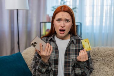 Evde genç bir kadın cep telefonundan kredi kartı bakiyesini kontrol ediyor kod hatası ya da sorun sonrası endişeli görünüyor. Kanepedeki beyaz saçlı kız başarısız alım satım, para kaybı ya da bloke edilmiş hesap yüzünden stresli.